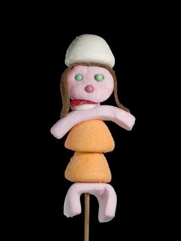 A female figurine made out of candies on a stick over a black background.