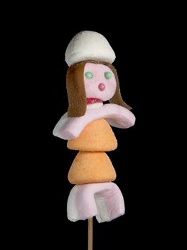 A female figurine made out of candies on a stick over a black background.