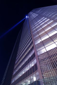  giant glass wall of modern skyscraper building on night sky background with blue light projector on the top