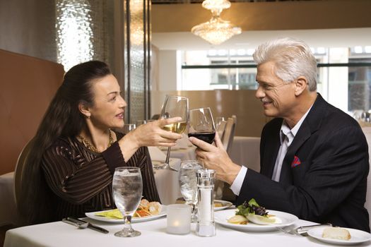 Caucasian mature adult male and prime adult female sitting at restaurant table toasting wine glasses.