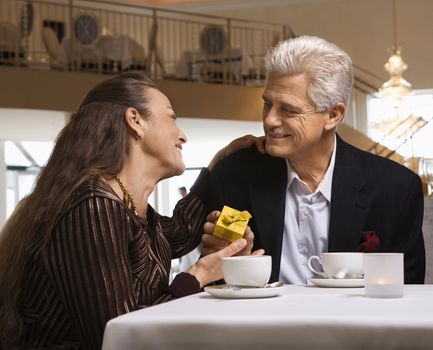 Caucasian mature adult male giving prime adult female gift box at restaurant table.