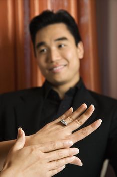 Prime adult Asian female admiring engagement ring with male in background.