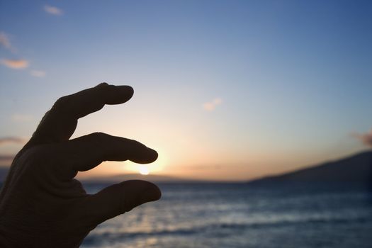 Hand in foreground with fingers around sunrise over the coast of Kihei, Maui, Hawaii, USA.