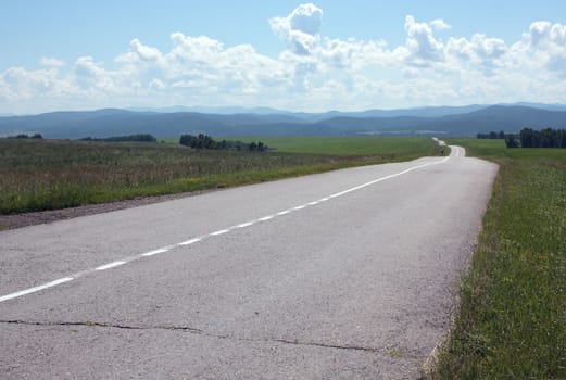 Landscape with the asphalted empty road, taking place waves on hills, clouds, the sky, mountains in distances. Beauty and calmness.