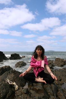 beautiful woman meditating on the rocks in ireland with her eyes closed, in the lotus position, showing a healthy way to live a happy and relaxed lifestyle in a world full of stress