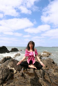 beautiful woman meditating on the rocks in ireland with her eyes closed, in the lotus position, showing a healthy way to live a happy and relaxed lifestyle in a world full of stress