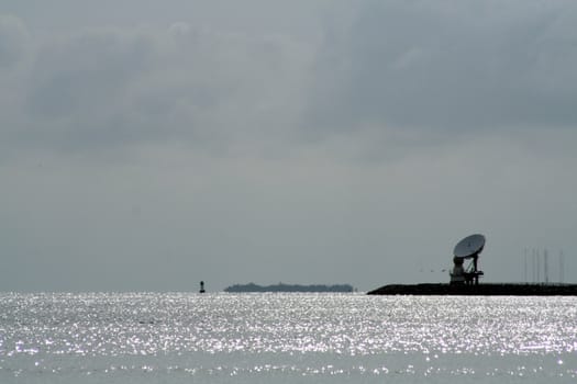 A satellite dish sitting on the ocean shore and pointed skyword.