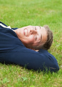 Young blond guy lying on grass, slit eyes because of bright sunlight