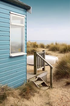 Viw of the side of a blue wooden beach hut with wooden terrace, looking towards the coast/beach. A white upturned boat rests in front of the hut amongst the sand and reed bushes.