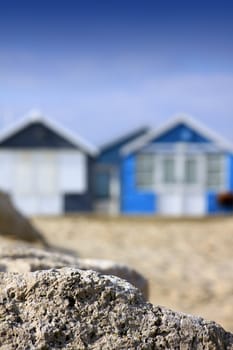 A portrait format image of blue painted wooden beach huts in soft focus to background, with sharp focus to a rock formation to front of image. Room for copy above image.