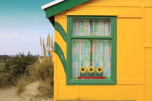 A landscape format image of the window of a brightly painted wooden beach hut. Located in Christchurch, Dorset Hampshire UK. Beach hut painted in yellow and green with wooden sunflowers inside its window.