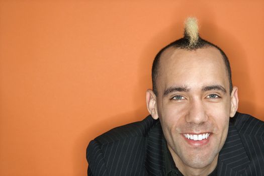 Head and shoulder portrait of Caucasian man in suit with mohawk smiling against orange background.