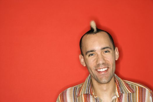 Caucasian man with mohawk smiling against red background.