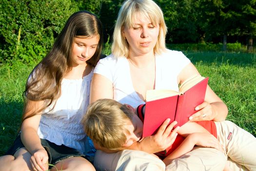 The young woman sits on a grass and reads the book to the children