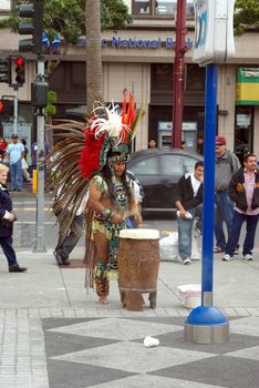 An Aztec street performer in full ceremonial dress plays a drum for dancers on a street corner in San Francisco California.