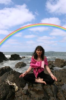 beautiful woman meditating on the rocks in ireland with her eyes closed, in the lotus position, showing a healthy way to live a happy and relaxed lifestyle in a world full of stress with a beautiful rainbow in the background