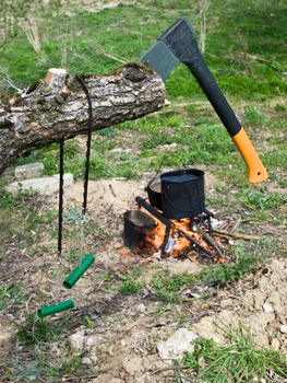 Hand saw and axe stuck in a tree, kettle with boiling water over campfire at background