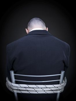 A businessman is tied up on a chair turning his back to the camera.