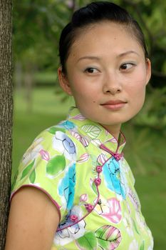 Chinese girl in park. Leaning on tree, kind looks, wearing traditional Chinese dress - qipao.