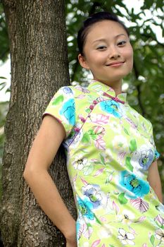 Chinese girl in park. Leaning on tree, kind looks, wearing traditional Chinese dress - qipao.