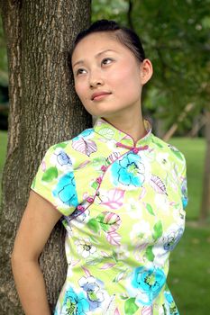 Chinese girl in park. Leaning on tree, kind looks, wearing traditional Chinese dress - qipao.