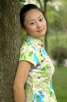 Chinese girl in park. Leaning on tree, kind looks, wearing traditional Chinese dress - qipao.