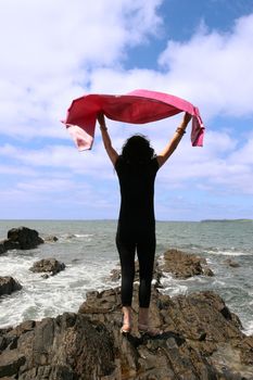 woman with high heels on the rocks at the waters edge happily waving a silk scarf in the air