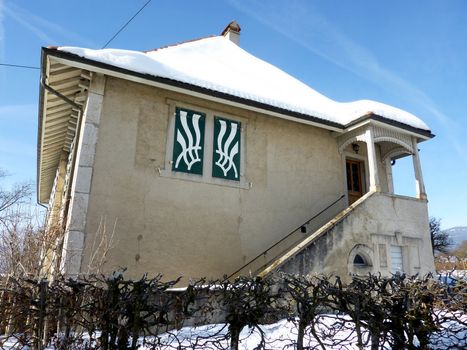 Grey house with snow on its roofs and and a hedge in front of it