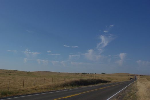 A 2 lane road cuts across prairie hills covered with dry grass with unusuale cloud formations in the background.