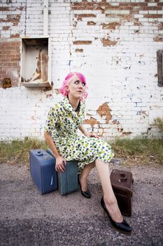 Woman with pink hair wearing polka dot dress in alley with suitcases