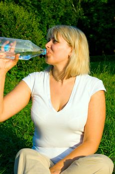 The woman-blonde drinks water from a plastic bottle against green trees