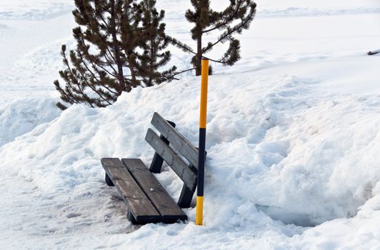 Bench amidst huge piles of snow with snow pole and bush