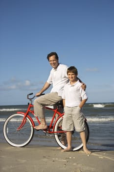 Caucasian father and pre-teen boy standing on beach with bicycle.