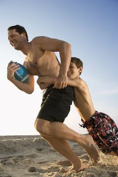 Caucasian mid-adult man running with football being tackled by pre-teen boy.