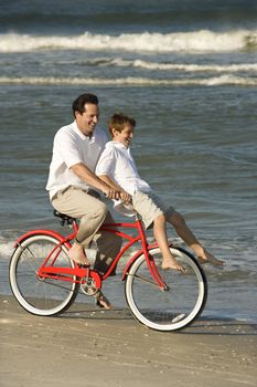 Caucasian mid-adult man riding bicycle on beach with pre-teen boy on handlebars.