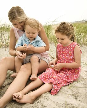 Caucasian mid-adult woman sitting with male toddler on lap and beside Caucasian female child on beach looking at shells.