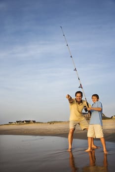 Caucasian mid-adult man shore fishing on beach with pre-teen boy and pointing.