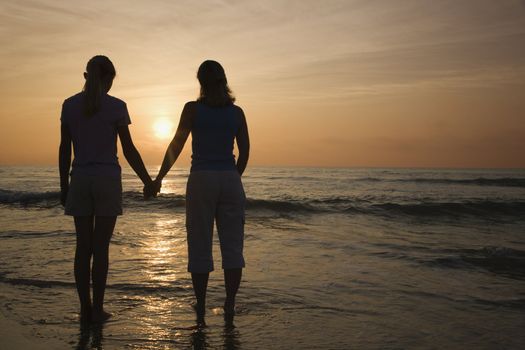 Caucasian mid-adult mother teenage and daughter standing on beach at sunset holding hands.