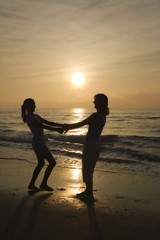 Caucasian mid-adult mother and teenage daughter holding hands spinngin on beach at sunset.