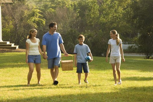 Caucasian family of four walking in park carrying picnic basket.