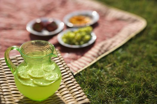 Green plastic pitcher of lemonade and lemons with a picnic spread out in the background.