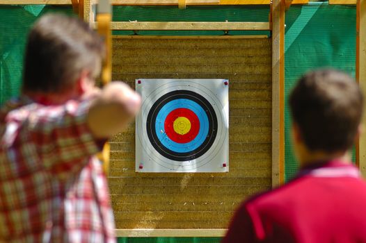 An archer aims at the bullseye of a distant target whilst a spectator looks on. Selective focus on the target.