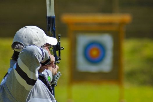 An archer aims at the bullseye of a distant target. Selective focus on the archer's head.