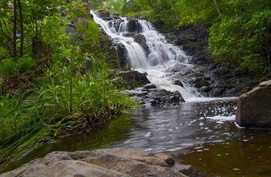 A waterfall falling into a pool surrounded by a green spring forest in Northern Minnesota.