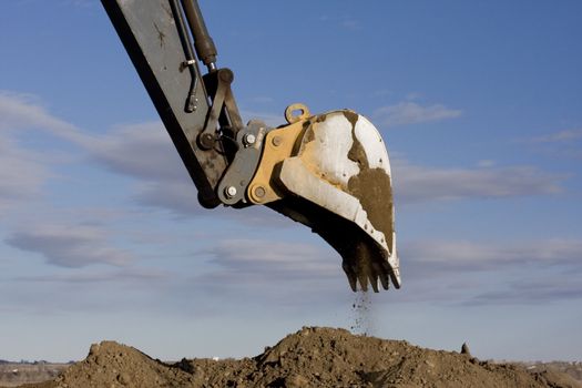 Excavator arm and scoop digging dirt at construction site against blue, partially cloudy, sky