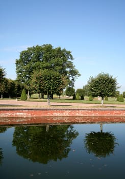 In the well-groomed park two trees are reflected in the calm pond
