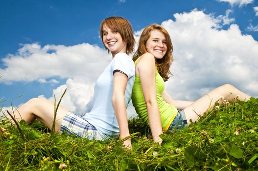 Two young teenage girl friends sitting back-to-back on summer meadow