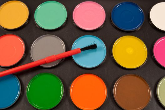 Close up of many dry, round, coloured watercolour paint tablets arranged over black with paintbrush.