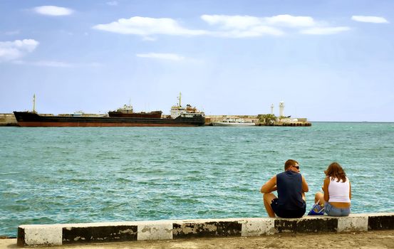 The tourists, a man and a woman sit at the berth and waiting for his ship. What would go on the tour. In the background a large transport ship and a lighthouse. City Yalta. The peninsula of Crimea. Ukraine.