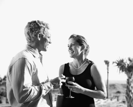 Black and white image of mid-adult Caucasian couple holding wine glasses and smiling at each other.
